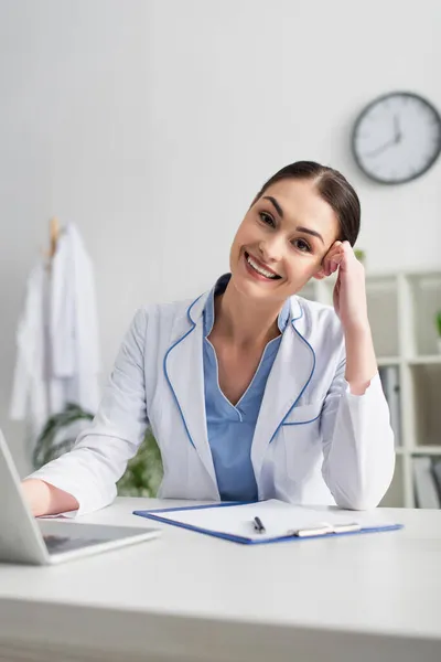 Médico positivo mirando la cámara cerca de la computadora portátil y portapapeles en la clínica - foto de stock