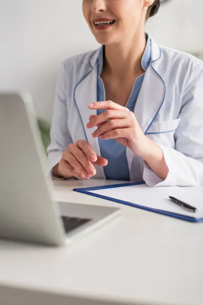 Vista cortada de médico sorrindo ter chamada de vídeo no laptop perto da área de transferência e caneta na clínica — Fotografia de Stock