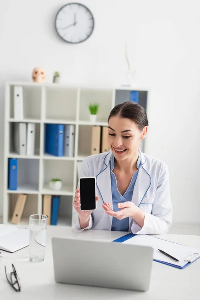 Médecin souriant tenant smartphone lors d'un appel vidéo sur ordinateur portable à l'hôpital — Photo de stock