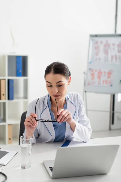 Doctor holding eyeglasses near laptop and glass of water in clinic — Stock Photo