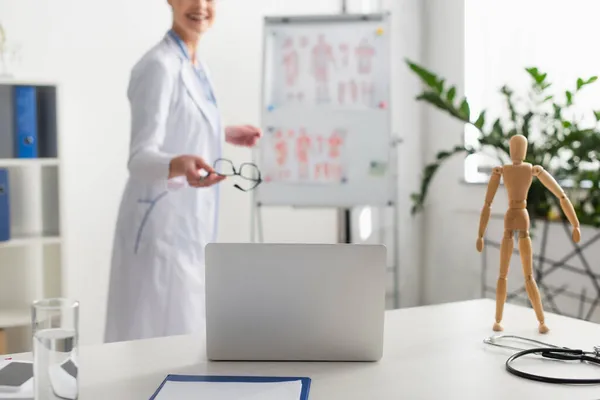 Vista recortada de la computadora portátil cerca del portapapeles y el médico borroso que sostiene las gafas en clink - foto de stock