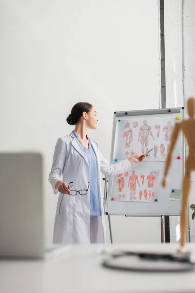 Side view of smiling doctor with eyeglasses pointing at flip chart near blurred laptop on table in hospital — Stock Photo