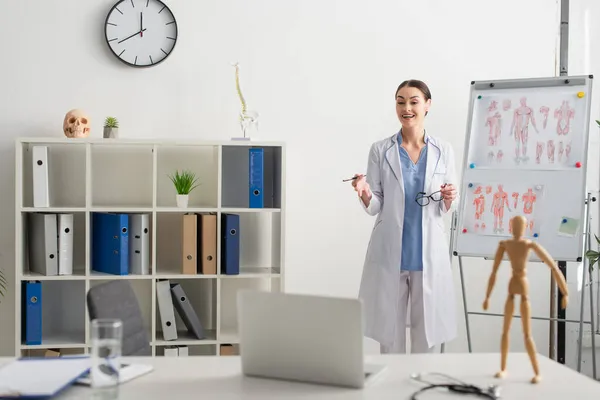 Smiling doctor holding eyeglasses near flip chart and looking at laptop in clinic — Stock Photo