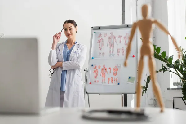 Médico morena sosteniendo pluma y anteojos cerca de rotafolio y portátil en la clínica - foto de stock