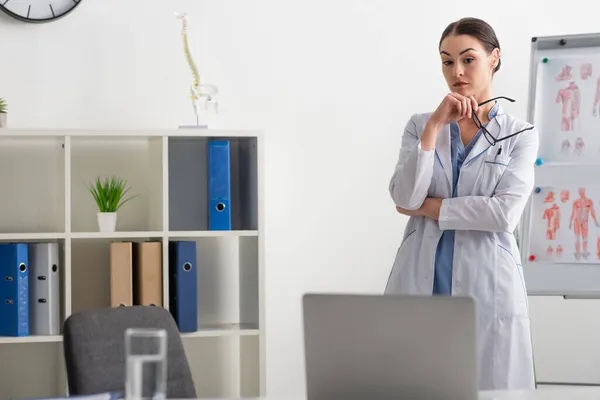 Médico pensativo segurando óculos enquanto olha para laptop borrado na clínica — Fotografia de Stock