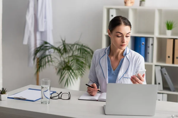 Doctor sosteniendo teléfono inteligente y pluma cerca de anteojos y vaso de agua en el escritorio - foto de stock