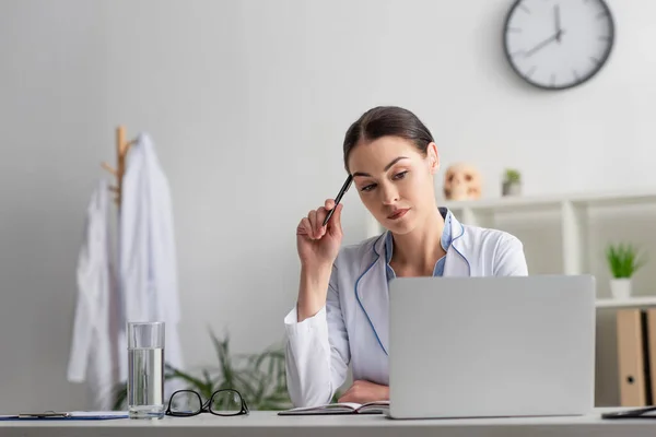 Médico reflexivo mirando el ordenador portátil mientras está sentado con la pluma en el lugar de trabajo - foto de stock