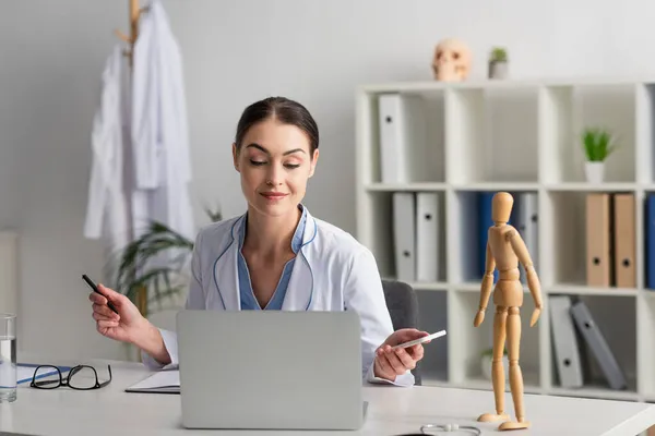 Medico che tiene smartphone e penna vicino al computer portatile e statuetta uomo in legno — Foto stock