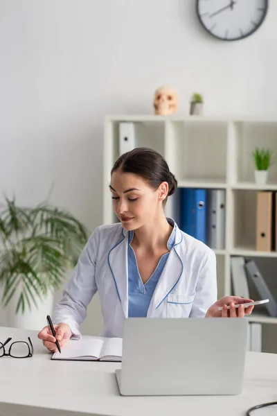 Medico bruna che tiene smartphone e scrive in notebook vicino a laptop offuscato — Foto stock