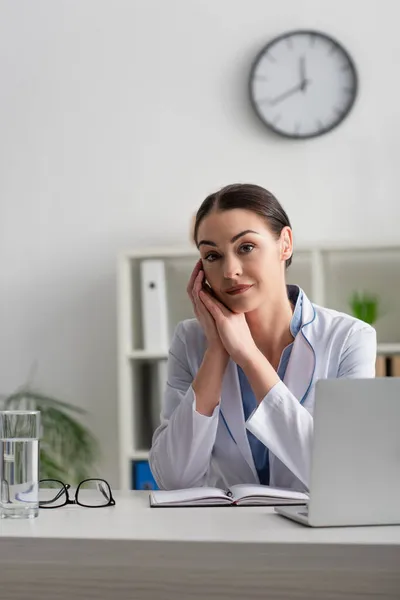 Morena médico mirando a la cámara mientras está sentado en el escritorio cerca de la computadora portátil, portátil y vaso de agua - foto de stock