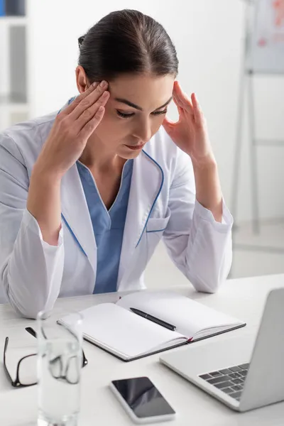 Morena médico tocando la cabeza mientras piensa cerca de dispositivos y portátil en el escritorio - foto de stock