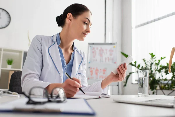 Médecin souriant tenant stylo et regardant smartphone tout en étant assis sur le lieu de travail dans le bureau — Photo de stock