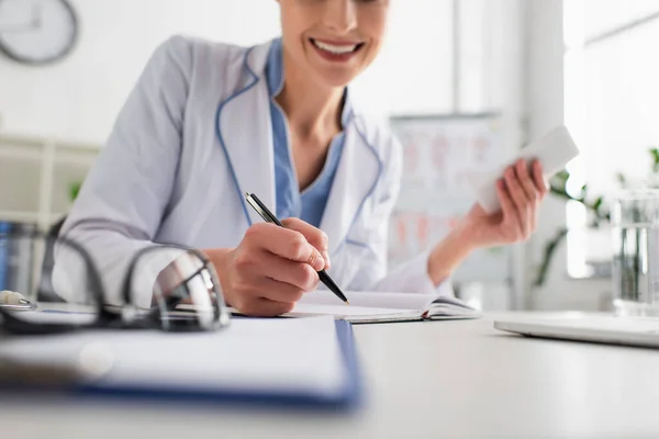 Cropped view of smiling doctor with smartphone writing in notebook on blurred foreground — Stock Photo