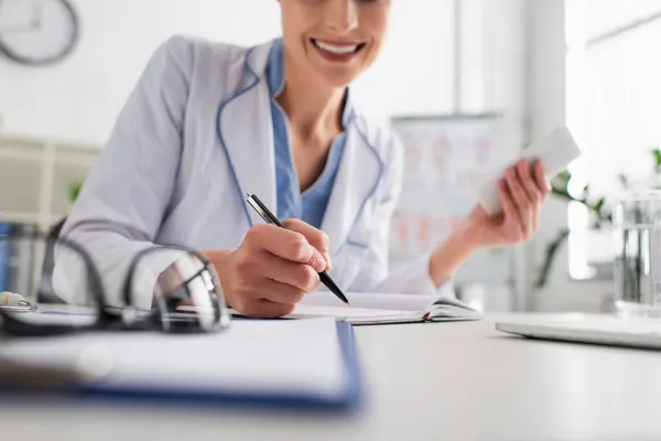 Ausgeschnittene Ansicht eines lächelnden Arztes mit Smartphone-Schrift in Notizbuch auf verschwommenem Vordergrund — Stockfoto