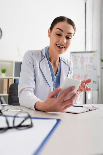 Smiling doctor gesturing during video call on blurred smartphone — Stock Photo