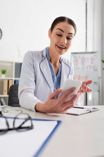 Medico sorridente che fa gesti durante la videochiamata su smartphone sfocato — Foto stock