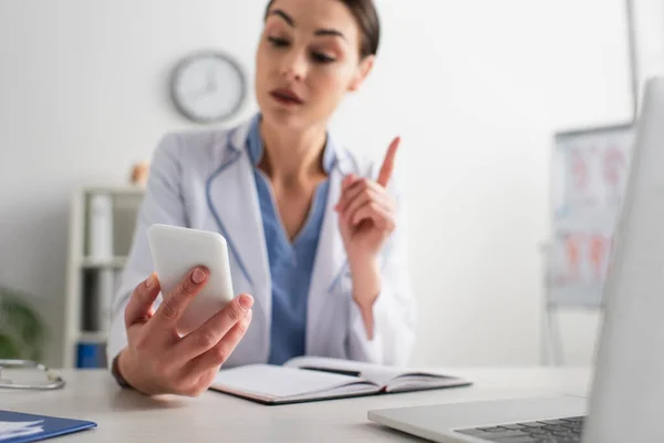 Blurred doctor pointing with pen while looking at mobile phone at workplace — Stock Photo