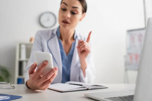 Médico borroso apuntando con pluma mientras mira el teléfono móvil en el lugar de trabajo - foto de stock