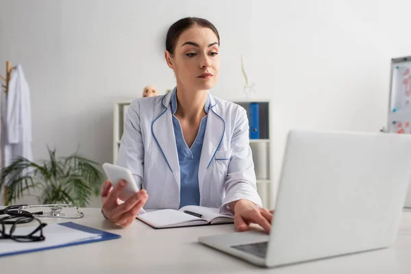 Brunette doctor holding cellphone while typing on blurred laptop in clinic — Stock Photo