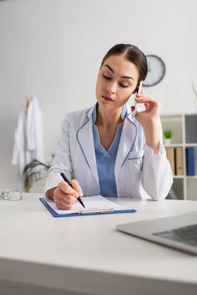 Médico falando no telefone celular enquanto escrevia na área de transferência perto do laptop borrado — Fotografia de Stock