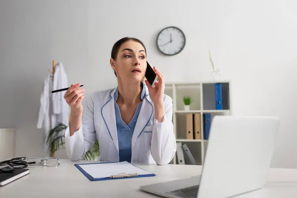 Doctor holding pen while talking on mobile phone near empty clipboard and blurred computer — Stock Photo