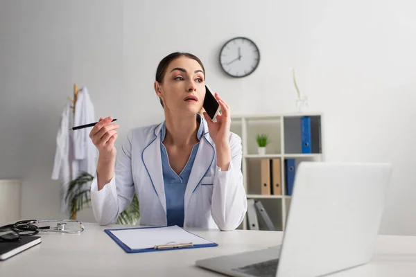 Médico segurando caneta enquanto fala no telefone celular perto da área de transferência vazia e computador borrado — Fotografia de Stock