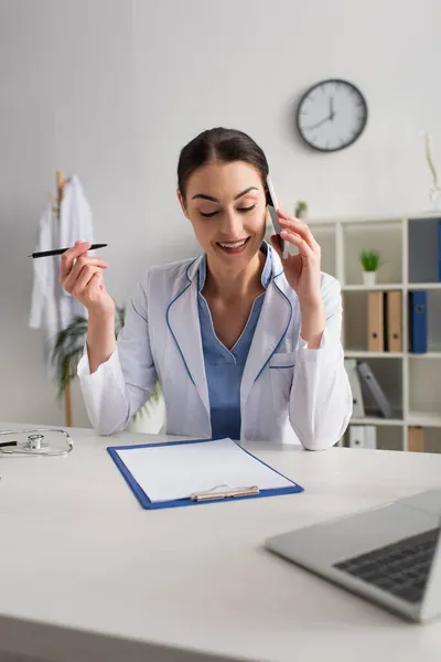 Médico feliz segurando caneta enquanto fala no smartphone perto da área de transferência vazia e laptop borrado — Fotografia de Stock