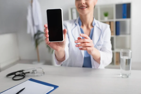 Doctor borroso mostrando teléfono inteligente con pantalla en blanco cerca de un vaso de agua y portapapeles en el escritorio - foto de stock