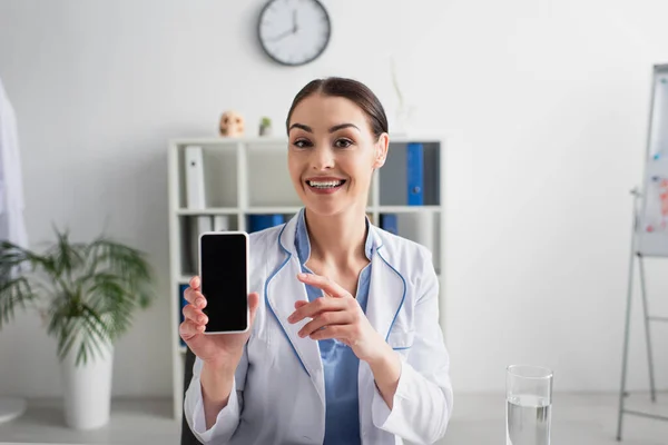 Щасливий брюнетка лікар вказує на мобільний телефон з порожнім екраном, дивлячись на камеру — стокове фото