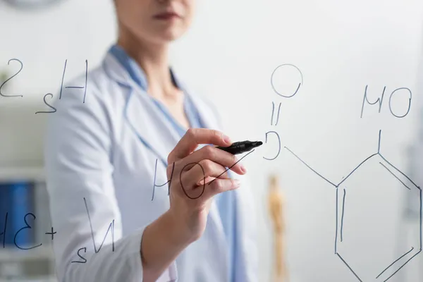 Partial view of blurred chemist with felt pen writing formulas on glass board — Stock Photo