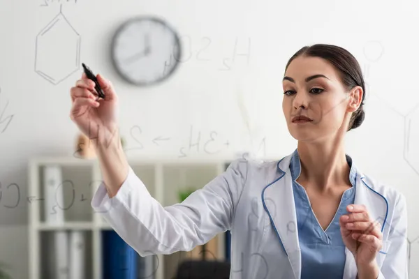 Brunette doctor in white coat writing chemical formulas on glass board in lab — Stock Photo