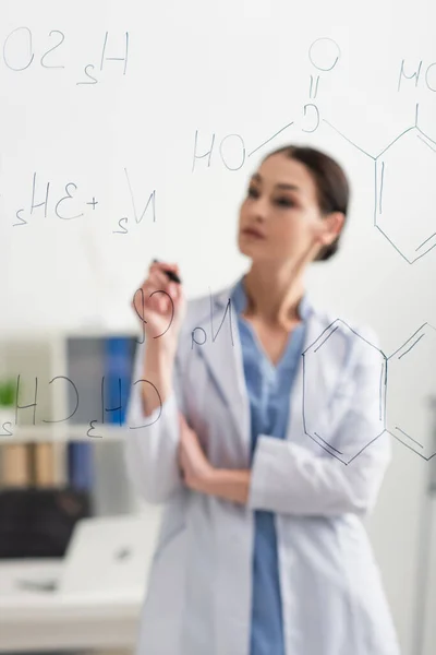 Blurred doctor in white coat standing near chemical formulas on glass board — Stock Photo