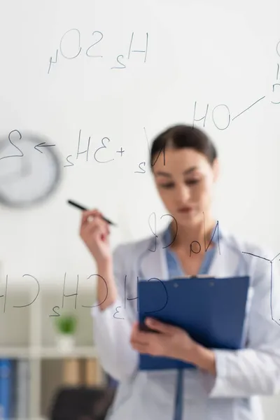 Selective focus of chemical formulas on glass board near scientist with clipboard and pen — Stock Photo