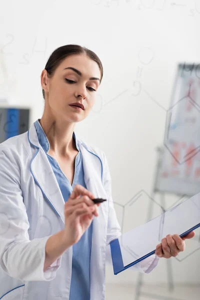 Brunette chemist holding clipboard while writing formulas on glass board — Stock Photo