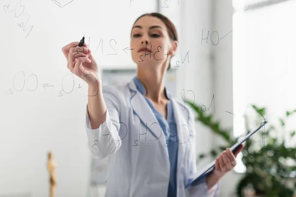 Doctor borroso con la pluma de fieltro y las fórmulas químicas de la escritura del portapapeles en tablero de cristal - foto de stock