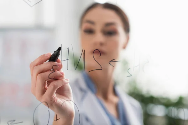 Blurred doctor with felt pen writing chemical formulas on glass board — Stock Photo