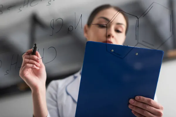 Blick in den niedrigen Winkel des unscharfen Chemikers, der Klemmbrett und Filzstift in der Nähe chemischer Formeln auf Glasplatte hält — Stockfoto