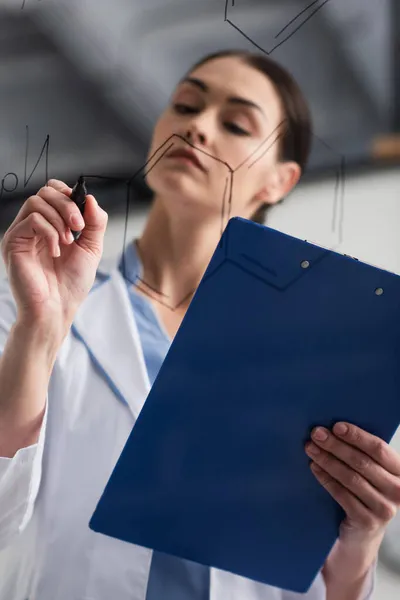 Vue à angle bas d'un scientifique flou avec presse-papiers écrivant des formules chimiques sur un panneau de verre — Photo de stock