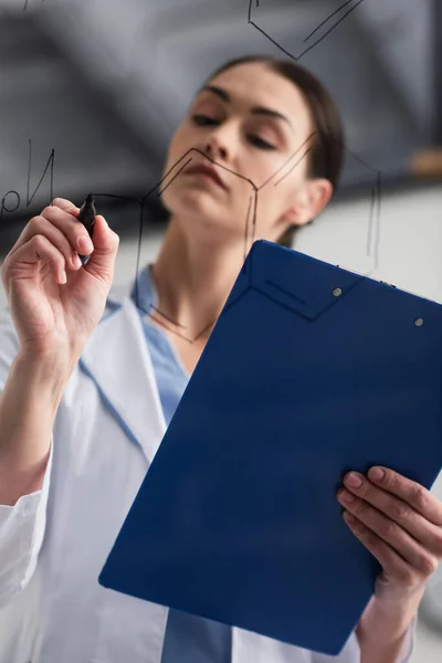 Low angle view of blurred scientist with clipboard writing chemical formulas on glass board — Stock Photo