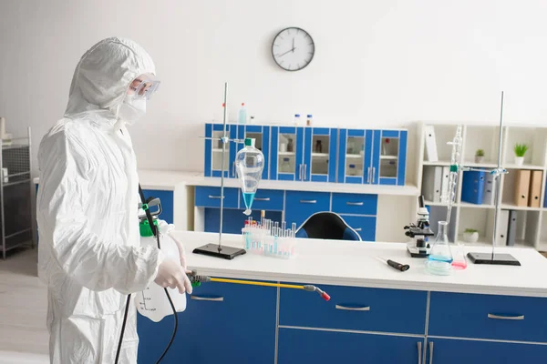 Médico en traje de materiales peligrosos haciendo desinfección del laboratorio cerca del equipo médico en el escritorio - foto de stock