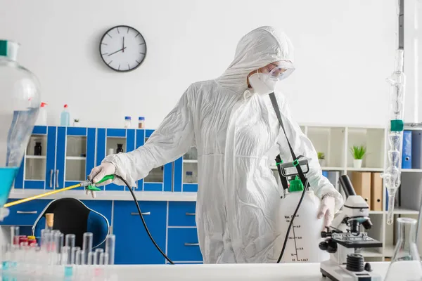 Científico en traje de materiales peligrosos haciendo desinfección del laboratorio cerca del microscopio y tubos de ensayo en el escritorio - foto de stock