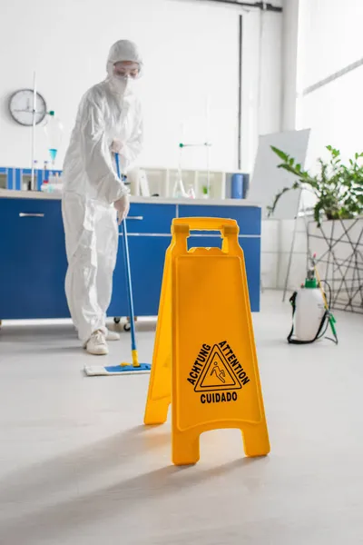 Placa de atenção perto do médico no chão de limpeza de terno hazmat com esfregão — Fotografia de Stock