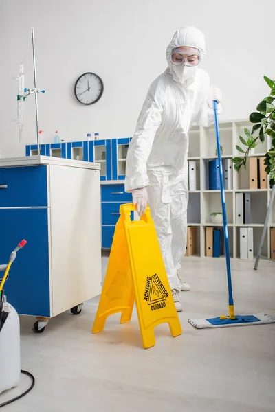 Doctor in hazmat suit holding mop and attention board while cleaning laboratory — Stock Photo