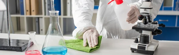 Vista recortada del médico en traje de materiales peligrosos que limpia el escritorio cerca de los frascos y microscopio, pancarta - foto de stock