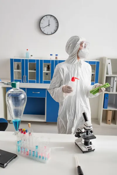 Doctor en traje de materiales peligrosos sosteniendo trapo y botella con detergente en el laboratorio - foto de stock