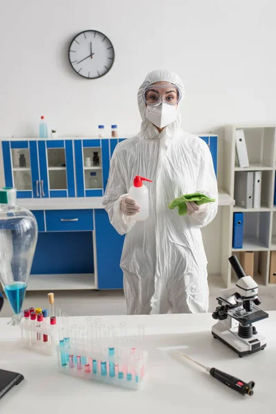 Científico en traje de materiales peligrosos sosteniendo la botella con detergente y trapo mientras mira la cámara - foto de stock