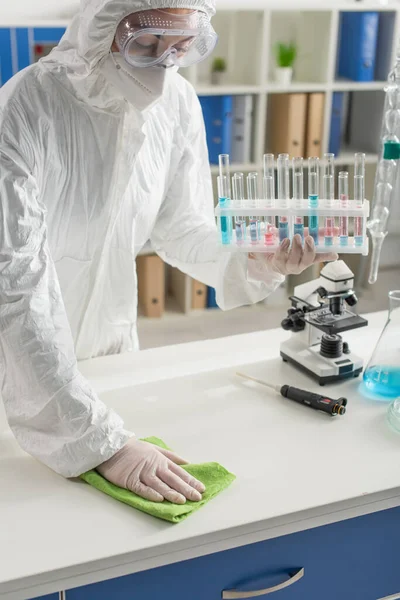 Médico em óculos e hazmat terno mesa de limpeza com pano enquanto segurando tubos de ensaio — Fotografia de Stock