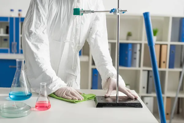 Vue partielle du médecin en combinaison Hazmat essuyant bureau tout en faisant la désinfection du laboratoire — Photo de stock