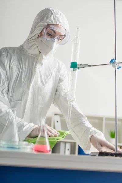 Científico en traje de materiales peligrosos y gafas que limpian el escritorio con trapo mientras hace la desinfección del laboratorio - foto de stock