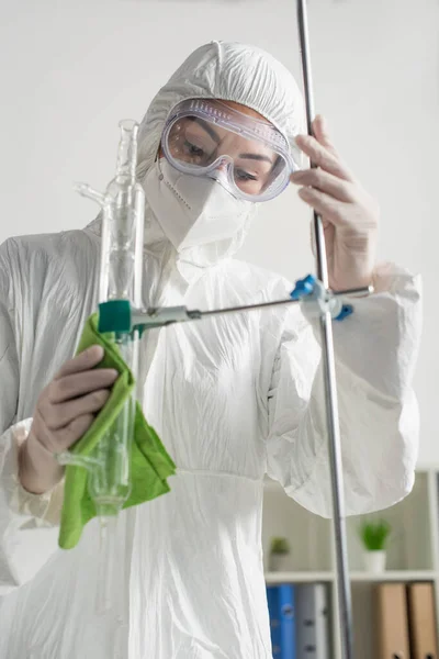 Mujer con gafas, máscara médica y traje de hazmat limpiando trípode en la clínica - foto de stock
